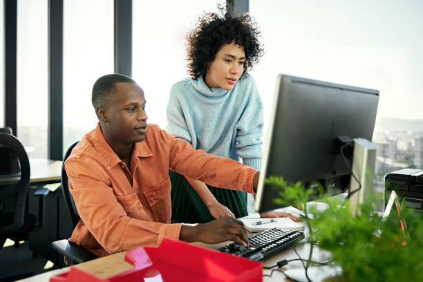 Young business partners working on project in modern office Waist-up view of casually dressed Black and Hispanic associates collaborating as they sit and stand at flexible workstation, looking at desktop PC monitor. black technology person stock pictures, royalty-free photos & images