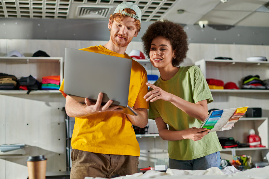 Smiling young african american craftswoman holding color swatches and pointing at laptop while standing near coffee to go and clothes in print studio, small business success concept